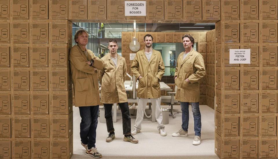 Remi Versteeg, Stijn de Weerd, Joep Dyckhoff en Pieter Kraaijenbrink in grijze jassen voor een Boring bureau in een wand van Boring dozen, OBJECT Rotterdam 2025